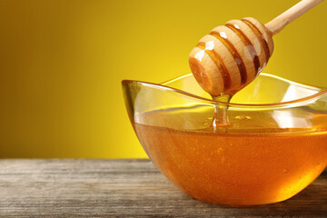 Delicious honey flowing down from dipper into bowl on wooden table against yellow background,...