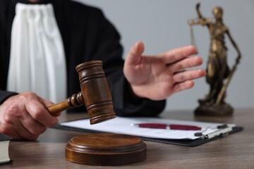 Judge with gavel and papers sitting at wooden table against light grey background, closeup