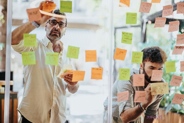 Different aged business people write business ideas on sticky notes over a glass wall,...