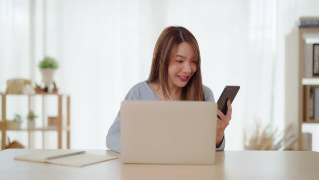 Young asian woman happy working on computer laptop and hand typing chat message on mobile smartphone at home. Video call, Online meeting