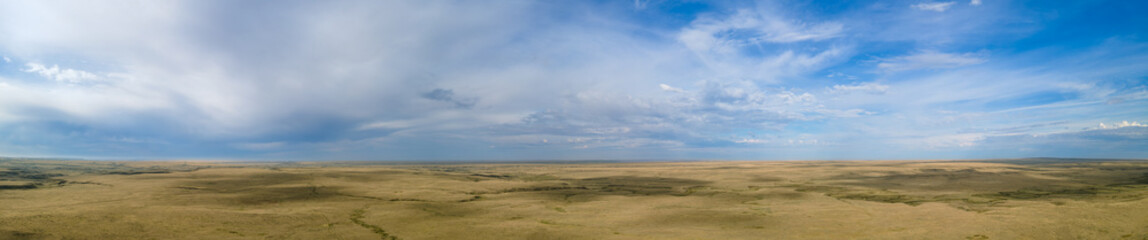 Aerial panorama of a vast flat dry prairie landscape with no trees under a sky with white to blueish colored clouds and patches of clear bright blue sky.
