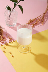 A glass of soy milk placed on pink and yellow surface with soybeans and a pot of green leaves. Soy...