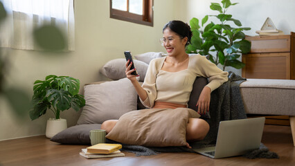 Pretty Asian woman is sitting on the floor beside her bed and using her smartphone, watching videos.