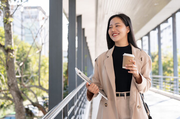 A beautiful businesswoman is holding a takeaway coffee cup and her laptop while walking on a skywalk