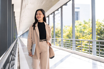 An attractive Asian businesswoman is holding her laptop while walking on a skywalk in the city.