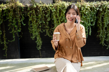A happy Asian woman is enjoying talking on the phone and sipping coffee while relaxing on a bench.