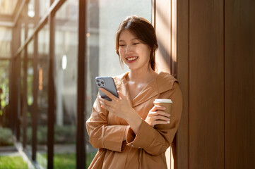 An attractive Asian woman is leaning on the wall outdoors, using her smartphone and sipping coffee.