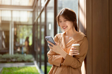 An attractive Asian woman is leaning on the wall outdoors, using her smartphone and sipping coffee.