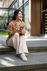 Fototapeta premium Beautiful Asian woman is sitting on the stairs with her phone and a takeaway coffee cup in her hands