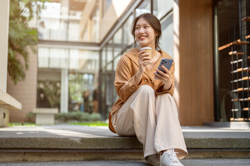 Beautiful Asian woman is sitting on the stairs with her phone and a takeaway coffee cup in her hands