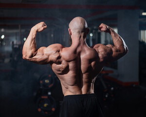 Muscular bald man posing shirtless. Bodybuilder demonstrating back muscles in the gym.