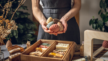 A woman soap maker holds soap in her hands in the form of colorful coniferous cones. A lot of different sliced pieces in a wooden box. Eco-friendly natural handmade cosmetics - obrazy, fototapety, plakaty