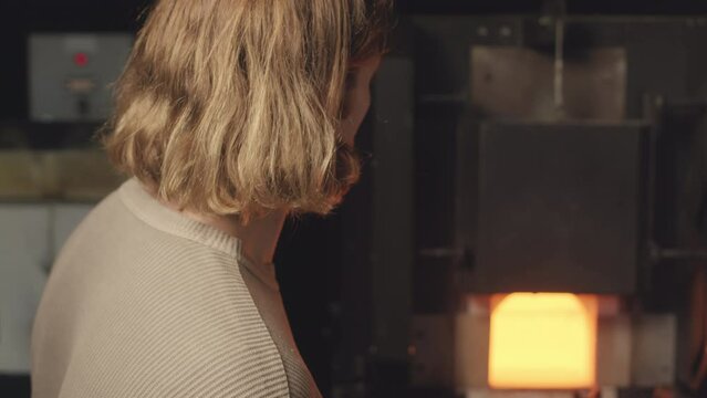 Tilting up slowmo portrait of young smiling Caucasian male gaffer posing for camera beside hot glass making oven at workshop