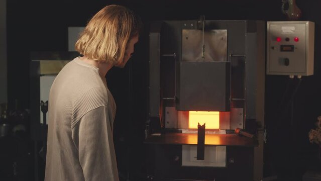 Medium slowmo portrait of young Caucasian male glassmaker looking at camera while standing by glass making oven at manufacturing factory