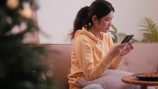 An Asian young woman is using a credit card to make online purchases on her smartphone while seated on the sofa in the cozy living room during the Christmas evening. Enjoy the convenience of holiday.