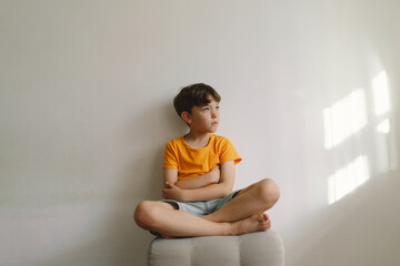 Obraz premium Cute boy sitting barefoot in a home. Beautiful light. Brunette boy 7 years old, casual clothes. Cozy home and lifestyle. Indoors.