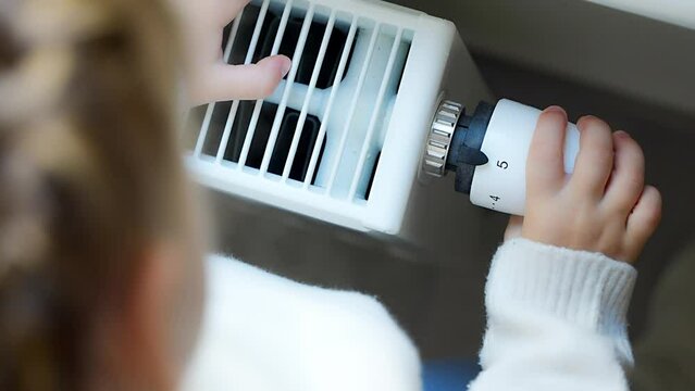Little girl in warm sweater and socks standing by window turning the battery heating knob and warming up from the heating radiator. Heating in an apartment, at home.