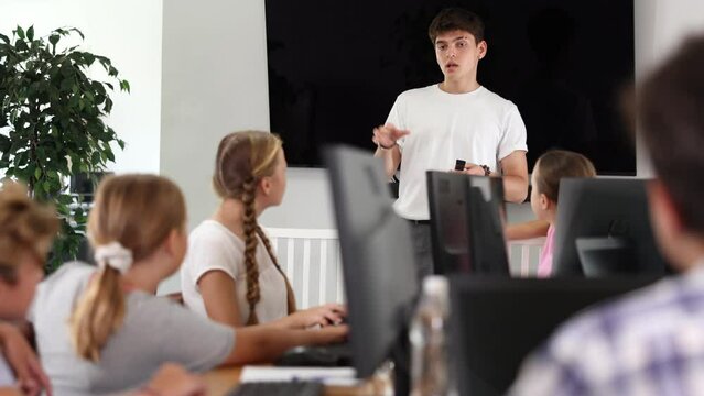 Young computer science teacher standing near an interactive whiteboard tells new material to teenage students in a school computer class