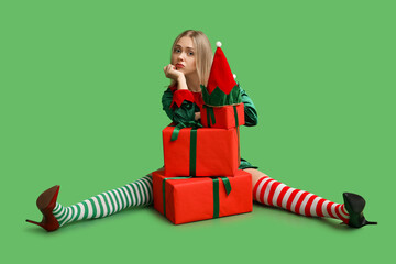 Sad young woman in elf costume with Christmas gift boxes on green background