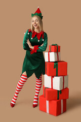 Beautiful young woman in elf costume with Christmas gift boxes on brown background