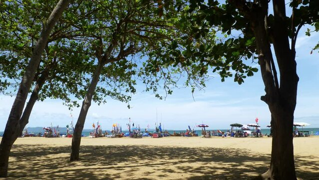 View of a beachfront with from under the shades of trees looking at colorful banners on wooden poles blown by the wind alongside beach umbrellas.