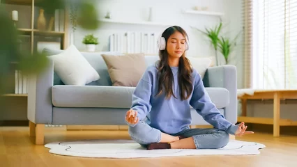 Fotobehang Happy young Asian woman practicing yoga and meditation at home sitting on floor in living room in lotus position and relaxing with closed eyes. Mindful meditation and wellbeing concept © Monster Ztudio