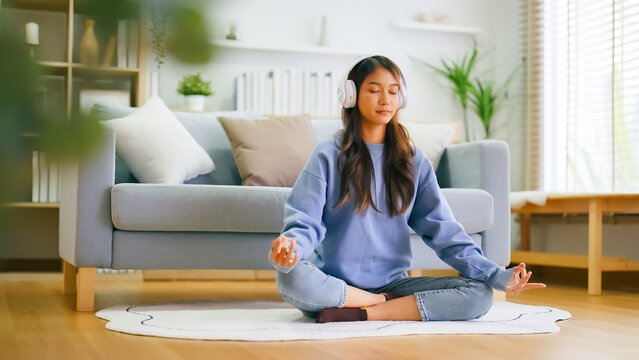Happy young Asian woman practicing yoga and meditation at home sitting on floor in living room in lotus position and relaxing with closed eyes. Mindful meditation and wellbeing concept