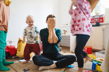 Stressed out mother sitting on floor while children running around her. - obrazy, fototapety, plakaty