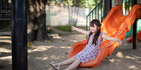 Smiling Asian little girl with fun sitting on slider at playground in the park. Education activity...