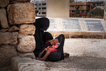 poor Muslim woman in a black hijab sits on the ground and holds her baby in her arms against the backdrop of a city in the Middle East. concept of poverty and misery. Internally displaced persons.