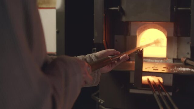 Cropped shot of unrecognizable male glassblower slowly rotating blowpipe with molten piece of glass on its end placed in burning stove with hot orange fire in it