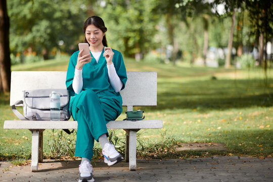 Cheerful medical nurse sitting on bench in park, eating lunch and scrolling through social media - Powered by Adobe