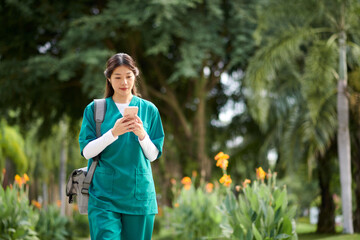 Young Vietnamese medical nurse checking notifications on smartphone when walking to hospital - obrazy, fototapety, plakaty