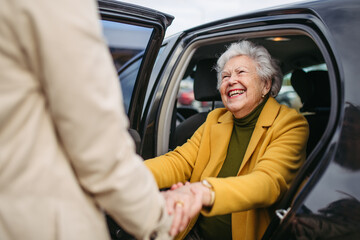 Senior lady getting out of the car, caregiver helping her, holding her hands. Elderly woman has problem with standing up from the car back seat. - obrazy, fototapety, plakaty