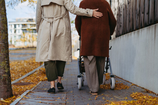Grandmother and mature granddaughter on a walk in city streets, during windy autumn day. Caregiver and senior lady with mobility walker enjoying the fall weather.