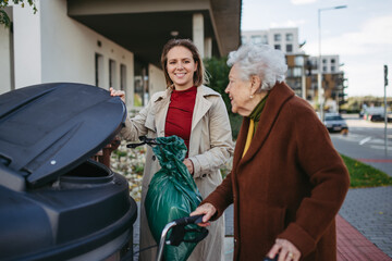 Woman helping elderly neighbor throw away trash into garbage can, waste container in front of their...