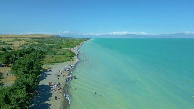 Aerial view of a large blue azure sea lake in clear sunny weather. People are swimming on the beach and pine trees are growing, boats are standing. Drone video, climbing up. Lake Sevan, Armenia.
