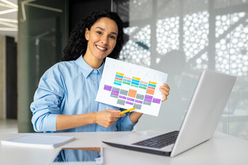 Cheerful hispanic woman sitting with laptop at table in modern office, talking on video call, showing graphs on webcam, conducting seminar, showing presentation, looking at camera.
