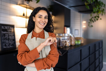 Portrait of a young beautiful Hindu woman at the workplace of a barista, a waitress, smiling and looking at the camera, an employee holding glasses in his hands is working inside a cafe restaurant.