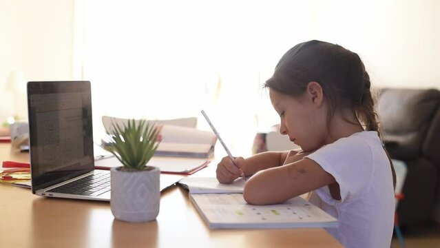 Child study at home. Girl in white t-shirt sit in table write note on copybook  and study with zoom online infront of laptop. Kid hold the hand on head with thoughtful face task exercise