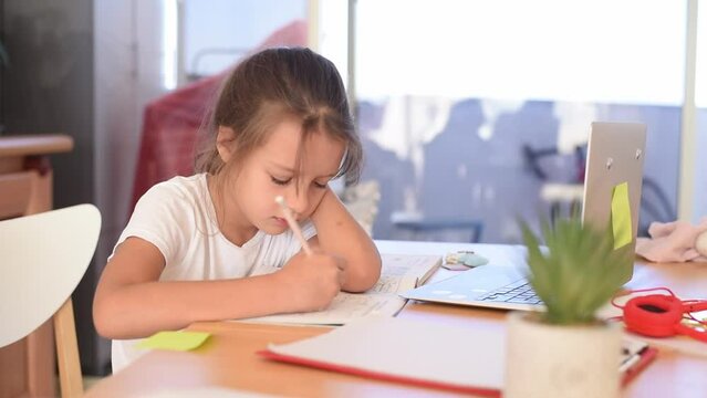 Child study at home. Girl in white t-shirt sit in table write note on copybook  and study with zoom online infront of laptop. Kid hold the hand on head with thoughtful face task exercise