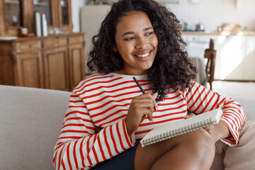 Pretty african american girl of 20s writing in her diary sitting on sofa in striped white and red clothes, noting down her feelings, emotions and thoughts, memories of last weekend with boyfriend - 677505612
