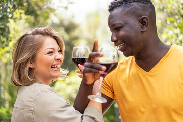 An interracial couple shares a toast with glasses of red wine, their laughter symbolizing a joyful...