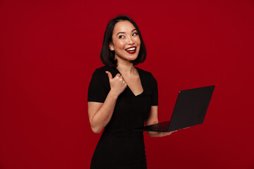 Cheerful woman working on laptop and showing thumbs up isolated over red wall