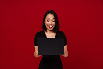 Fototapeta premium Excited happy asian woman using laptop isolated over red wall