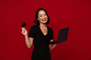 Naklejka premium Joyful woman using laptop computer and credit card for online shopping isolated over red wall