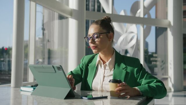 Stylish businesswoman preparing for marketing meeting and drinking coffee in large office hall USA. Successful beautiful Art Director starting online meeting and waving hello to front camera on tablet