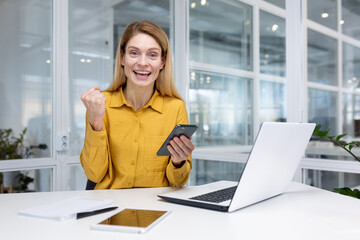 Happy blonde business woman sitting at a desk in a bright office, holding a smartphone in her hand, enjoying winning the lottery, reading good news, getting a promotion, looking at camera.