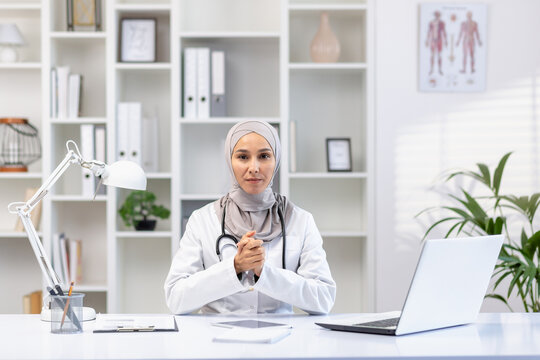 Portrait of serious asian muslim female doctor in hijab sitting at table in hospital office with stethoscope around neck, arms folded, looking at camera.
