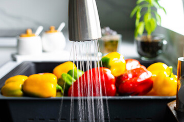 Yellow and red pepper are washed in a special tray for vegetables. The stream of water pours on the pepper in the sink. The interior of a modern kitchen